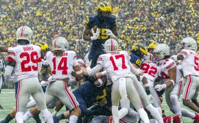 Michigan running back Hassan Haskins leaping over Ohio State players during a football game.